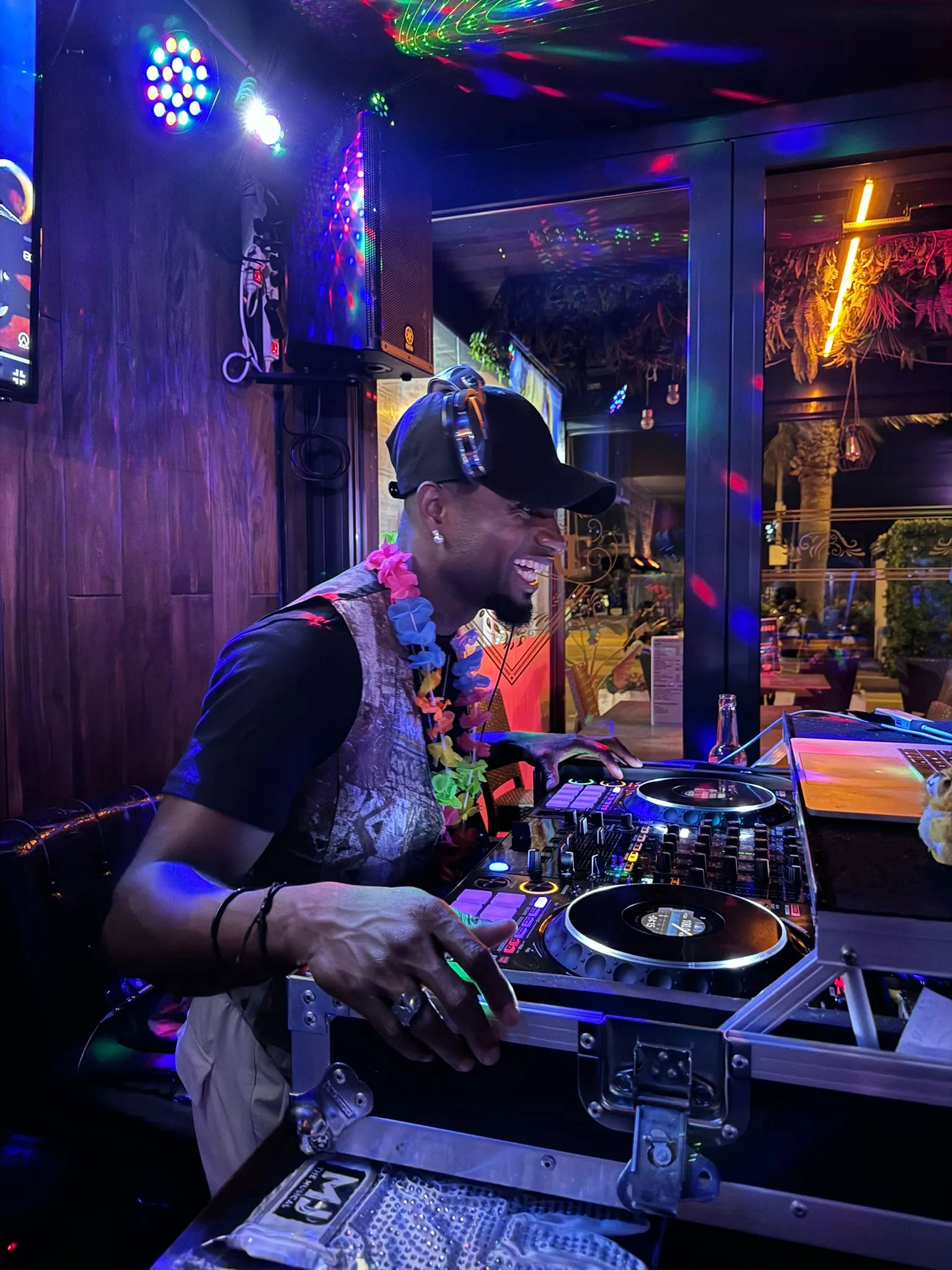DJ aux platines lors d'une soirée, casque autour du cou et collier de fleurs, ambiance colorée
