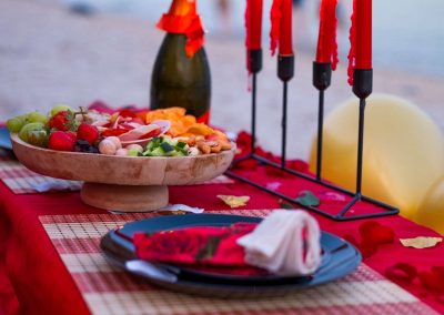 Table dressée sur la plage au coucher du soleil, bougies rouges, champagne et plateau de mets