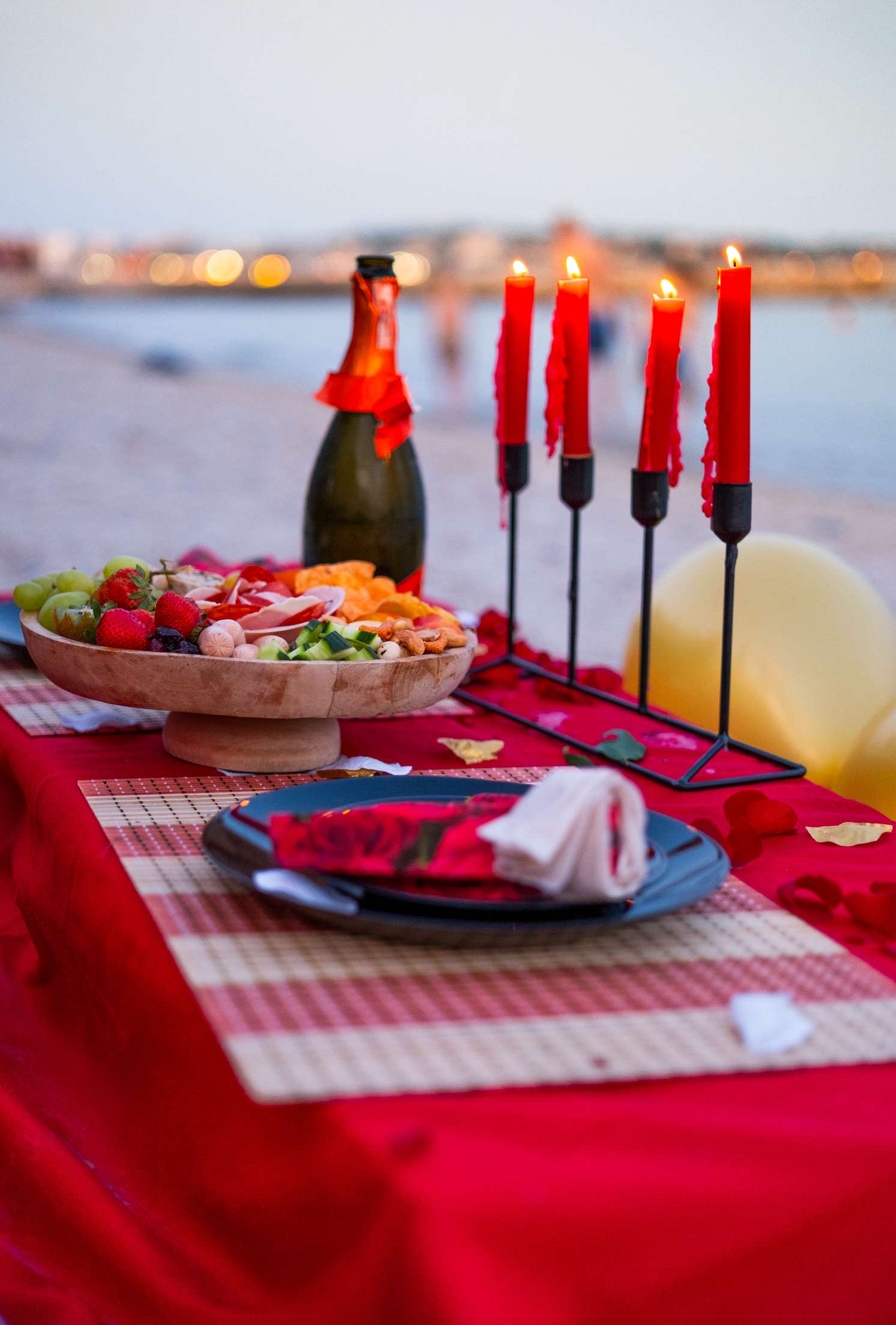Table dressée sur la plage au coucher du soleil, bougies rouges, champagne et plateau de mets