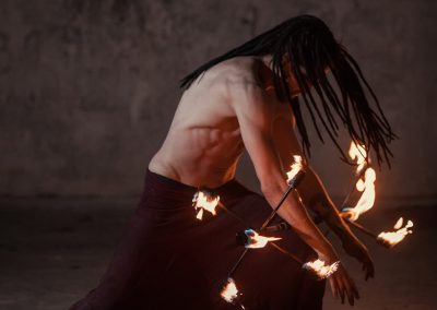 Performeur de feu torse nu, accroupi, manipulant des torches enflammées dans un décor sombre
