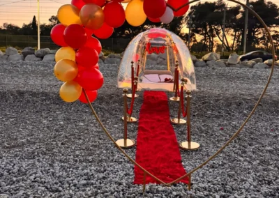 Demande en mariage sur la plage : bulle transparente illuminée, arche de cœur en ballons et tapis rouge au coucher du soleil