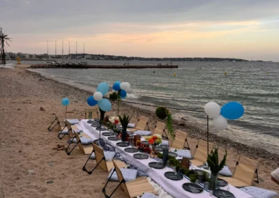 Table basse dressée sur le sable face à la mer, ballons bleus et guirlandes lumineuses au coucher du soleil